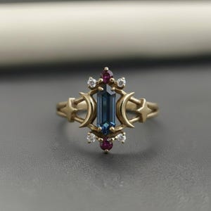 May include: A gold-colored ring featuring a rectangular, blue gemstone at its center. The ring is adorned with crescent moon and star motifs, along with small, round, red and white gemstones. The ring has a delicate, celestial design.