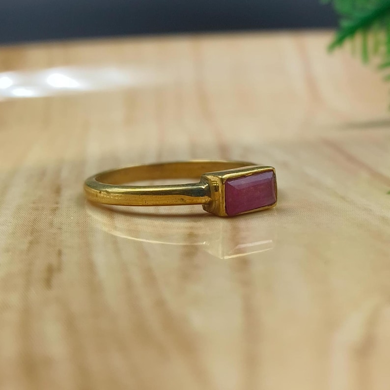 Pu&ograve; includere: Un anello color oro con una gemma rettangolare rosa-rossastra. L'anello &egrave; posto su una superficie chiara. La gemma &egrave; il punto focale, con la fascia dorata che fornisce uno sfondo contrastante. L'anello &egrave; un gioiello.