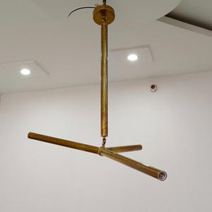 Puede incluir: Un candelabro moderno de color latón, con una varilla vertical central y tres brazos horizontales. La lámpara está suspendida del techo y presenta un diseño minimalista. Este accesorio de iluminación es una adición elegante a cualquier hogar.