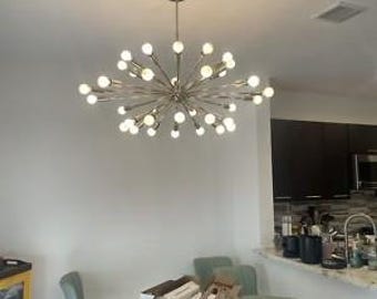 May include: A multi-arm sputnik chandelier with a metallic finish and numerous small, round, glowing white light bulbs extending outward from a central hub, suspended from the ceiling above a dining area with a wooden table and chairs.