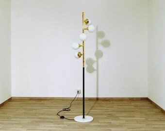 May include: A modern floor lamp with a black and gold metal frame. The lamp features six white globe-shaped lights and a white marble base. The lamp is plugged into a wall outlet and is positioned in a room with hardwood flooring.