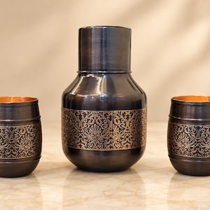 May include: A dark metallic pitcher and two matching tumblers featuring a copper-toned intricate floral pattern around the center. The vessels have a smooth, dark finish with a visible copper interior rim, showcasing a consistent design across all pieces.