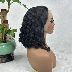May include: A black, curly wig styled in a bob cut, displayed on a mannequin head. The wig features a lace front and is styled with loose, natural-looking curls. The mannequin has a fair skin tone and is set against a white background.