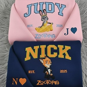 Puede incluir: Dos sudaderas, una rosa y otra azul marino, con diseños bordados. La sudadera rosa dice "JUDY" con un personaje de conejo y el texto "ZOOTOPIA". La sudadera azul marino dice "NICK" con un personaje de zorro y el texto "ZOOTOPIA".