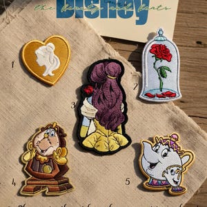 Puede incluir: Parches bordados con personajes y elementos de La Bella y la Bestia de Disney. Los diseños incluyen una silueta de corazón, una rosa en una cúpula de cristal, Bella, Din Don y la Sra. Potts. El texto de la imagen dice "Disney" y "the beauty and beats".