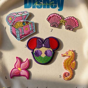 May include: Embroidered patches featuring Disney-themed designs. Includes a pink treasure chest, two pink seashells with a starfish, a Minnie Mouse-inspired design with Ariel's tail, a pink flower, and a yellow seahorse. The word "Disney" is above the patches.