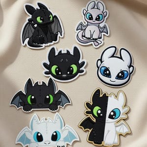 Peut inclure: Plusieurs écussons brodés représentant des dragons de dessins animés. Les écussons présentent des dragons noirs et blancs, avec des yeux verts et bleus. Les écussons ont des ailes et sont sur un fond beige.