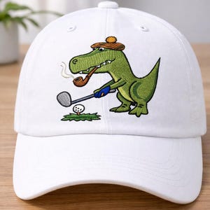 Pode incluir: Boné de beisebol branco com um dinossauro verde bordado jogando golfe. O dinossauro usa um chapéu e fuma um cachimbo. O taco de golfe está prestes a acertar a bola em um pedaço de grama verde.