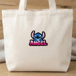 May include: A cream-colored tote bag with a cartoon patch of a blue alien with the word "ANGEL" in pink and black lettering. The bag has two white handles and is made of a canvas-like material.
