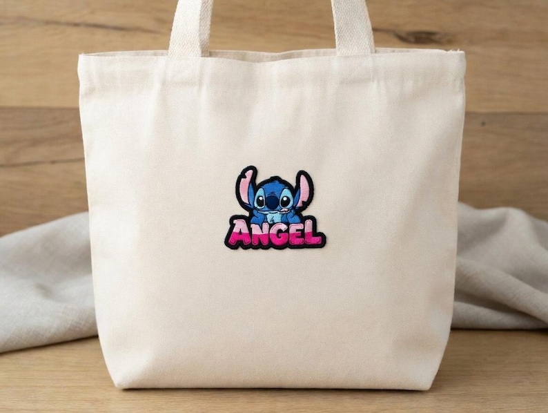 May include: A cream-colored tote bag with a cartoon patch of a blue alien with the word "ANGEL" in pink and black lettering. The bag has two white handles and is made of a canvas-like material.