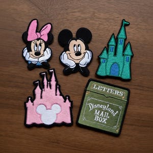 May include: Embroidered patches featuring Mickey Mouse, Minnie Mouse, and two castle designs. One patch is a green castle, and the other is pink. A green patch reads "Disneyland Mail Box" and "Letters".