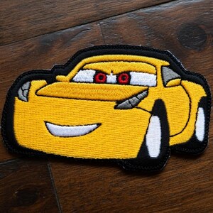 May include: Embroidered patch of a yellow cartoon race car with black edging. The car has white wheels, red eyes, and a smiling mouth. The patch is on a dark wooden surface.