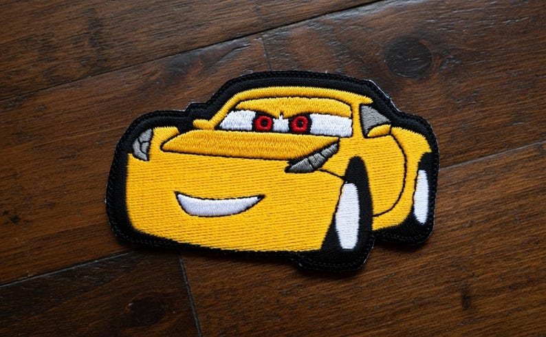 May include: Embroidered patch of a yellow cartoon race car with black edging. The car has white wheels, red eyes, and a smiling mouth. The patch is on a dark wooden surface.