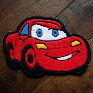 May include: Embroidered patch of a red race car with blue eyes and a white mouth, smiling. The car has black tyres and a black outline. Yellow accents are on the headlights. The patch is on a wooden surface.