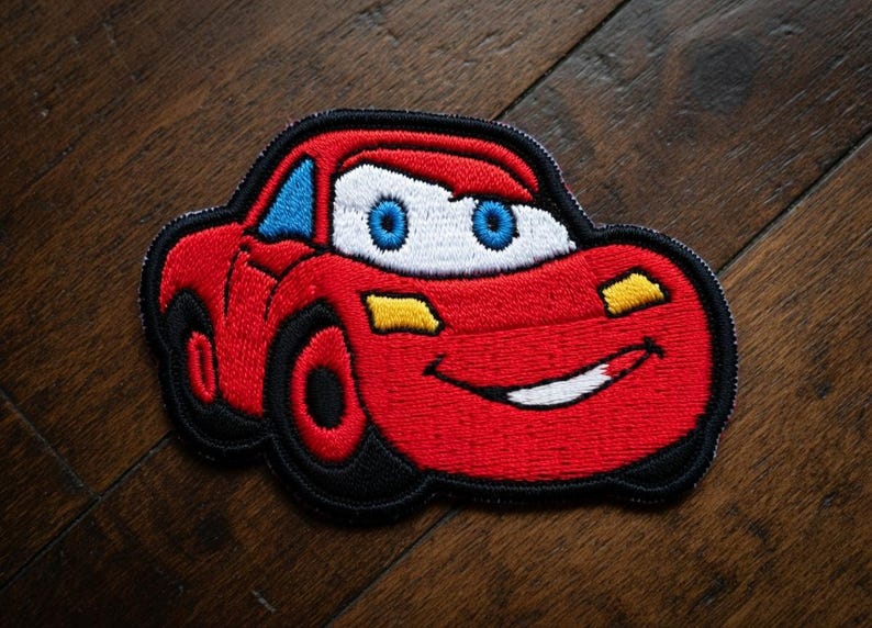 May include: Embroidered patch of a red race car with blue eyes and a white mouth, smiling. The car has black tyres and a black outline. Yellow accents are on the headlights. The patch is on a wooden surface.