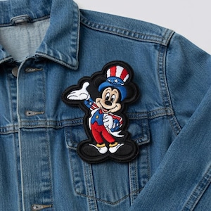 May include: A denim jacket with a Mickey Mouse patch. The patch features Mickey in patriotic attire, including a red, white, and blue hat and outfit. The patch is black with a cloud-like border.