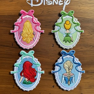 以下が含まれることがあります： 刺繍されたディズニーのプリンセスデザインのパッチ。各パッチは、オーロラ、ティンカーベル、アリエル、エルサなど、異なるプリンセスを楕円形のフレーム内に表示しています。ディズニーのロゴが上部にあります。パッチは1〜4の番号が付けられています。
