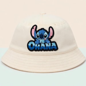 May include: Cream-colored bucket hat featuring a blue and black patch of the character Stitch with the word "OHANA" in blue lettering. The hat has a rounded crown and a downward-sloping brim, suitable for sun protection.