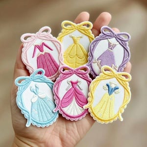Puede incluir: Parches bordados con vestidos de princesa coloridos en rosa, amarillo, azul y morado. Cada parche tiene un lazo en la parte superior y un marco decorativo. Los parches se sostienen en una mano.