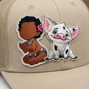 May include: Tan trucker hat featuring embroidered patches of a cartoon baby and a pig. The baby has brown skin, black hair, and a pink flower. The pig is white with black spots and pink ears and snout.