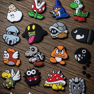 Pode incluir: Uma coleção de patches bordados coloridos com personagens de um videogame popular. Os patches incluem várias formas e designs, como fantasmas, tartarugas, cogumelos e estrelas. Os patches estão em uma superfície de madeira.