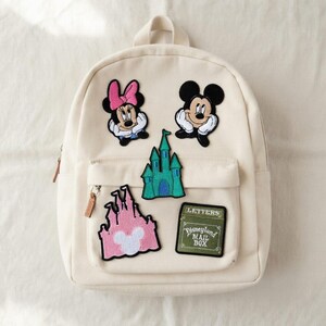 May include: Off-white backpack with multiple Disney-themed patches. Patches include Minnie Mouse, Mickey Mouse, a green castle, a pink castle, and a green patch that says "Disneyland Mail Box". The backpack has a front pocket and a top handle.
