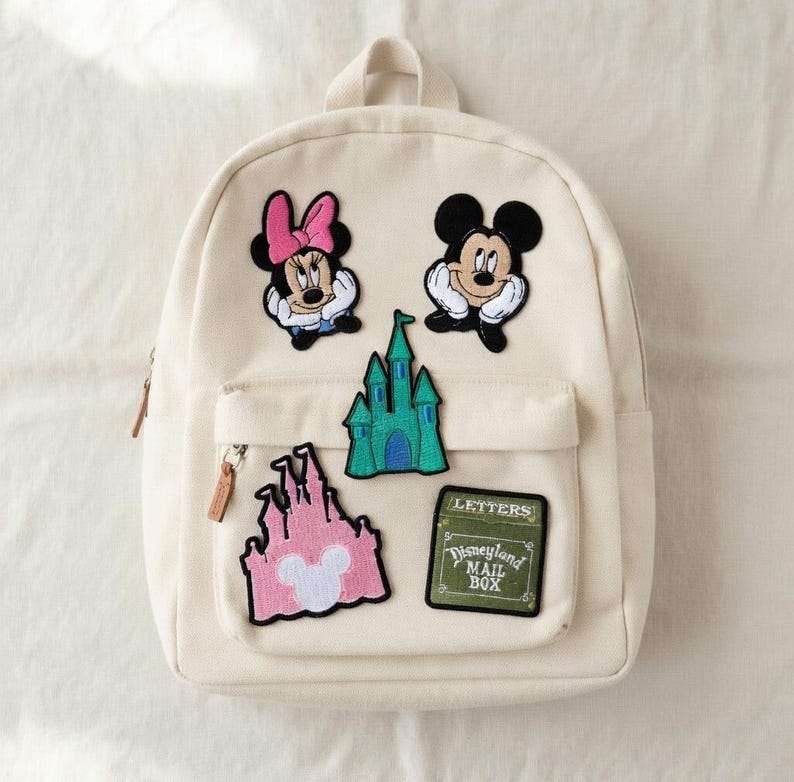 May include: Off-white backpack with multiple Disney-themed patches. Patches include Minnie Mouse, Mickey Mouse, a green castle, a pink castle, and a green patch that says "Disneyland Mail Box". The backpack has a front pocket and a top handle.
