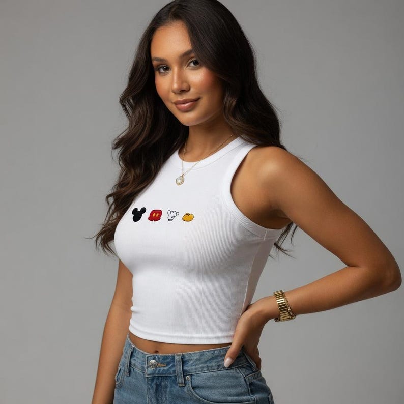 May include: White ribbed crop top with a crew neck and a colourful Mickey Mouse graphic. The graphic features Mickey's head, gloves, and shoes. The top is paired with blue jeans.
