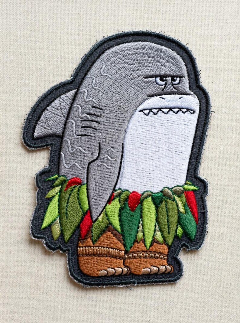 May include: Embroidered patch featuring a grumpy shark wearing a grass skirt and wooden sandals. The shark is gray with a white belly and a stern expression. The patch has a black border and is on a beige background.