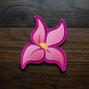 May include: Embroidered pink flower patch with a yellow center and a dark pink outline. The flower has five petals and is set against a dark wood grain background. This decorative patch can be ironed on or sewn onto fabric.