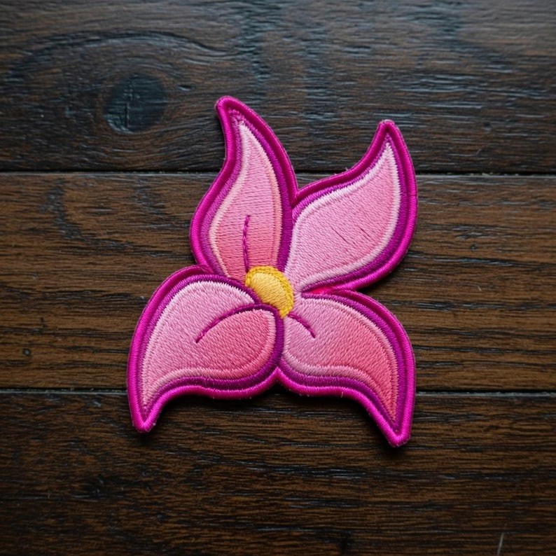May include: Embroidered pink flower patch with a yellow center and a dark pink outline. The flower has five petals and is set against a dark wood grain background. This decorative patch can be ironed on or sewn onto fabric.