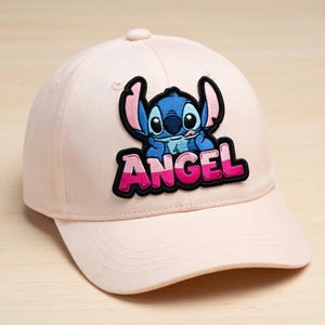May include: A light pink baseball cap featuring a patch with a blue cartoon character and the word "ANGEL" in pink and white. The cap has a curved brim and a button on top.