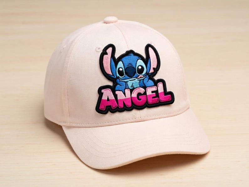 May include: A light pink baseball cap featuring a patch with a blue cartoon character and the word "ANGEL" in pink and white. The cap has a curved brim and a button on top.