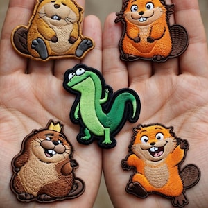 Könnte beinhalten: Fünf gestickte Aufnäher mit Zeichentricktieren. Es gibt drei Biber in Braun- und Orangetönen, eine grüne Eidechse und einen Biber mit Krone. Die Aufnäher haben detaillierte Nähte und sind vor einem neutralen Hintergrund platziert.