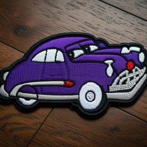 May include: Embroidered patch of a purple cartoon car with white accents, black outline, and a red detail. The car has a smiling face and white wheels. The patch is on a wooden surface.