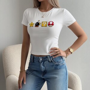 May include: White crop top with short sleeves, adorned with Super Mario game graphics: a star, a bomb, a question mark block, and a mushroom. The top is worn with blue jeans. The top is a casual, everyday wear item.