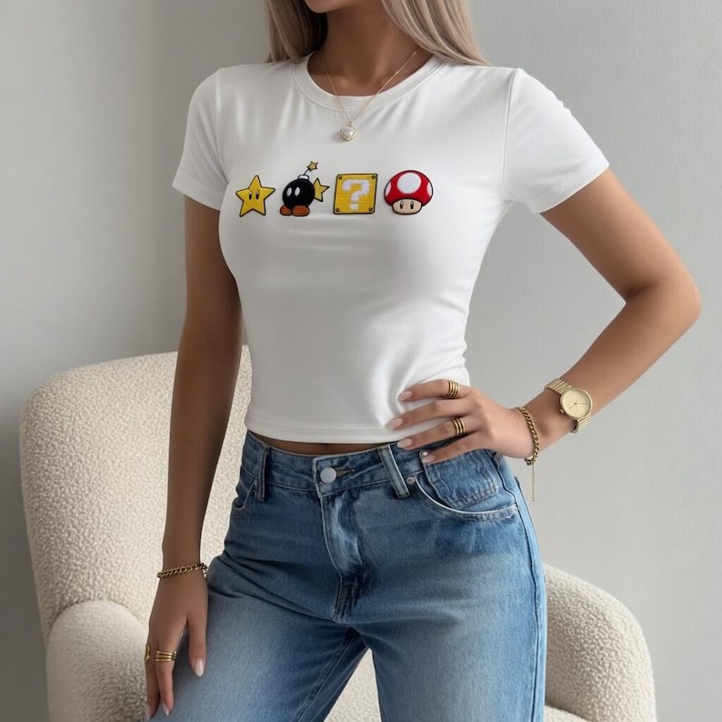 May include: White crop top with short sleeves, adorned with Super Mario game graphics: a star, a bomb, a question mark block, and a mushroom. The top is worn with blue jeans. The top is a casual, everyday wear item.