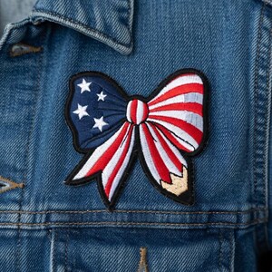 May include: A close-up of a denim jacket with a patriotic bow patch. The patch features a blue section with white stars and a red and white striped bow design. The jacket has a classic collar and a front pocket.