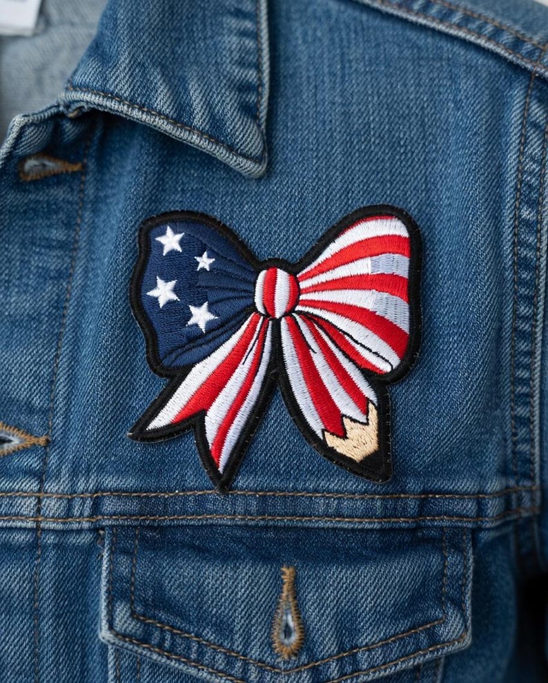 May include: A close-up of a denim jacket with a patriotic bow patch. The patch features a blue section with white stars and a red and white striped bow design. The jacket has a classic collar and a front pocket.