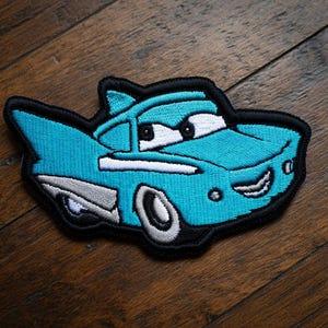 May include: Embroidered patch depicting a cartoon car in turquoise, white, and grey, with a black outline. The car has large eyes and a smiling mouth. This patch is designed for application on clothing or accessories.