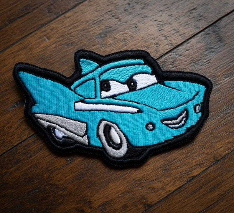 May include: Embroidered patch depicting a cartoon car in turquoise, white, and grey, with a black outline. The car has large eyes and a smiling mouth. This patch is designed for application on clothing or accessories.