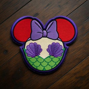May include: Embroidered patch in the shape of Mickey Mouse ears, featuring a mermaid design. The ears are red, outlined in purple. The center has a purple bow, two purple seashells, and green scales. The patch is on a dark wood surface.