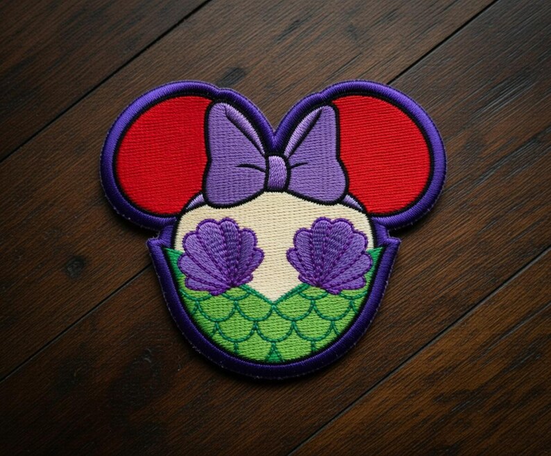 May include: Embroidered patch in the shape of Mickey Mouse ears, featuring a mermaid design. The ears are red, outlined in purple. The center has a purple bow, two purple seashells, and green scales. The patch is on a dark wood surface.