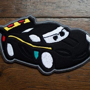 May include: Black embroidered car patch with white, yellow, red, and teal details. The patch features a cartoon car design with a grey border. The car has white headlights and a white windscreen.