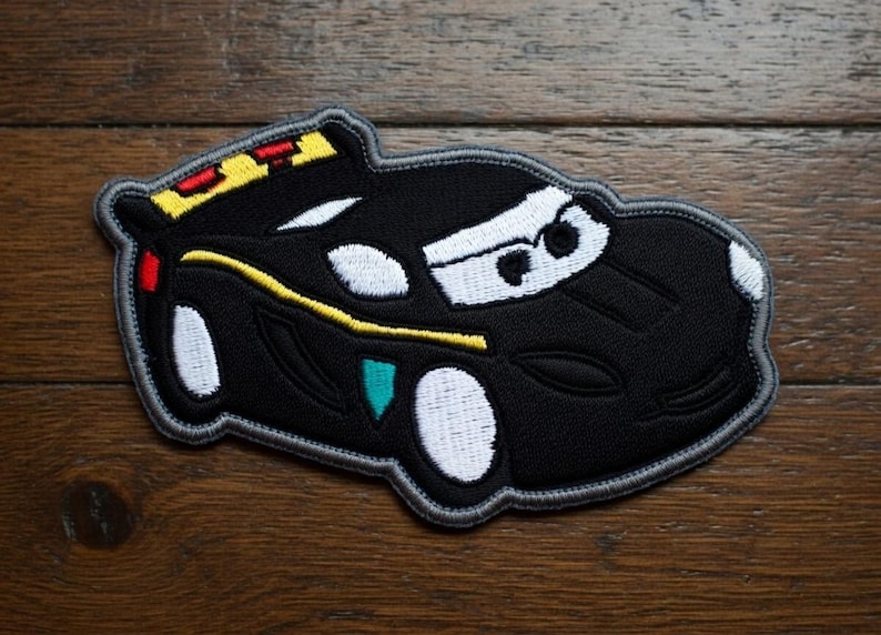 May include: Black embroidered car patch with white, yellow, red, and teal details. The patch features a cartoon car design with a grey border. The car has white headlights and a white windscreen.