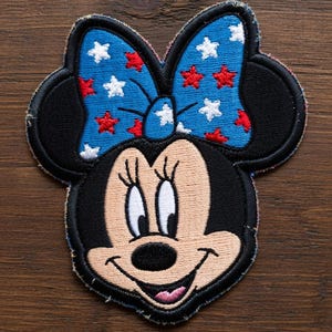 May include: Embroidered patch featuring Minnie Mouse's face. The patch has a black outline, black ears, and a blue bow adorned with red and white stars. Minnie's face is tan with a black nose and a smiling expression.