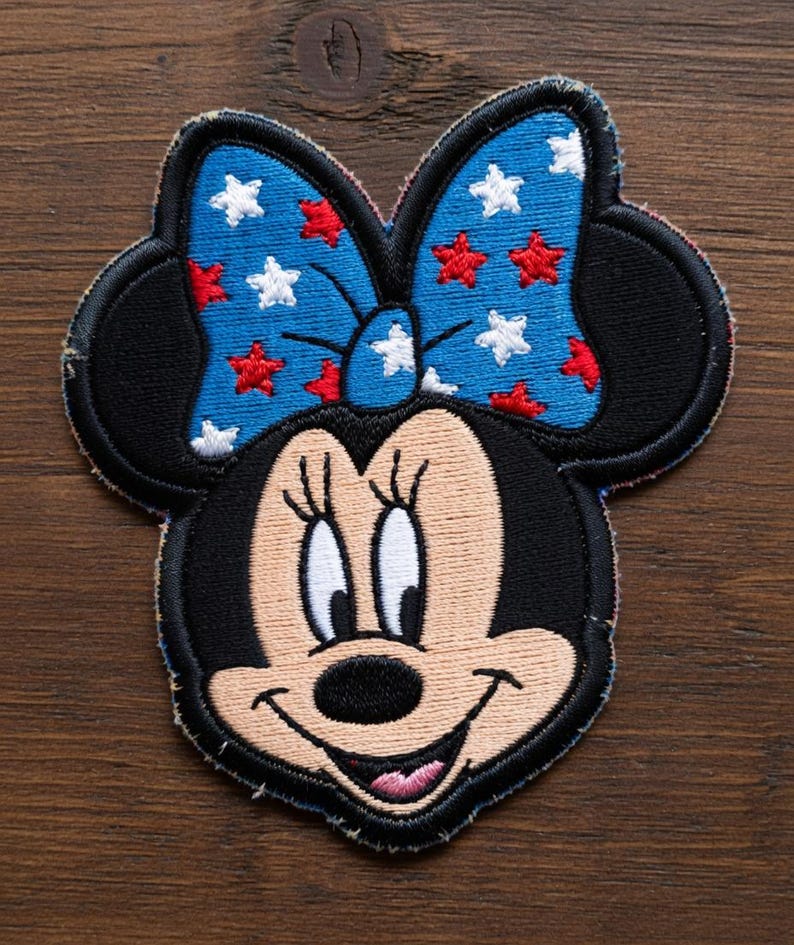 May include: Embroidered patch featuring Minnie Mouse's face. The patch has a black outline, black ears, and a blue bow adorned with red and white stars. Minnie's face is tan with a black nose and a smiling expression.
