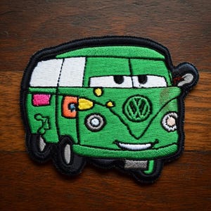 May include: Embroidered patch of a cartoon green van with a smiling face, black tyres, and white windows. The patch has a black border and includes the Volkswagen logo. The van has a pink and orange design on the side.