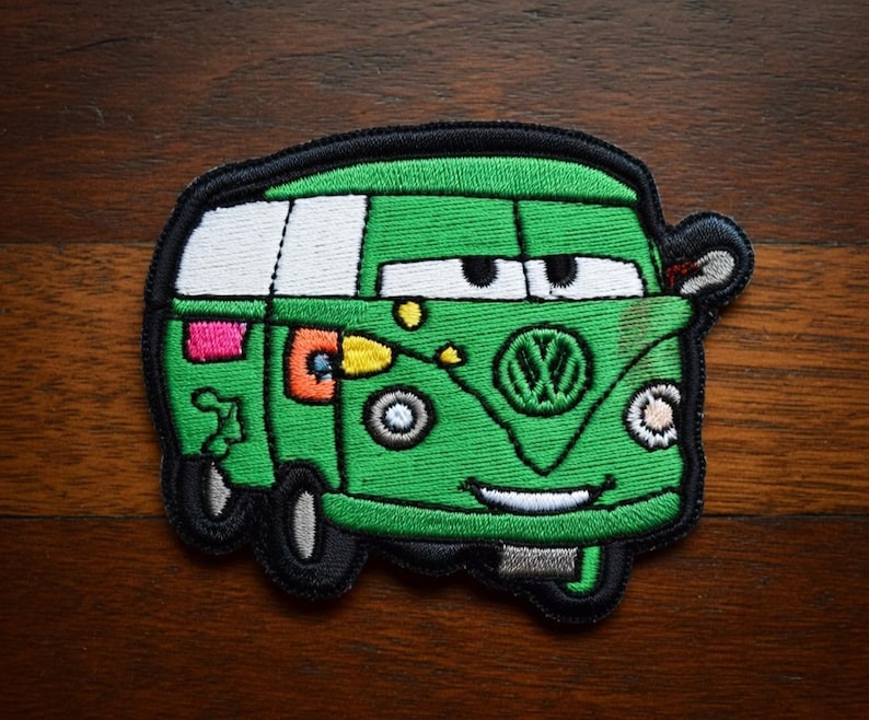 May include: Embroidered patch of a cartoon green van with a smiling face, black tyres, and white windows. The patch has a black border and includes the Volkswagen logo. The van has a pink and orange design on the side.