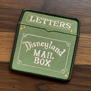 May include: Green embroidered patch with the words "LETTERS" and "Disneyland MAIL BOX" in white and gold lettering. The patch has a black border and is rectangular in shape.